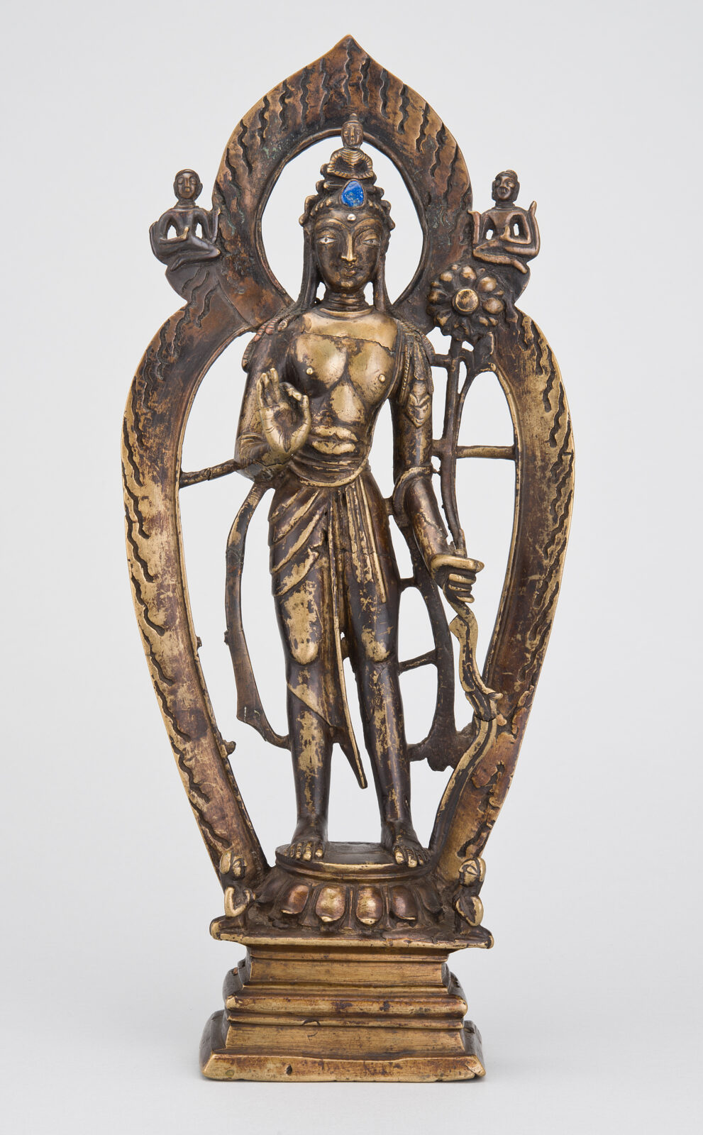 Bronze statue of a standing bodhisattva with a blue gemstone in the crown, holding a lotus stem, framed by an arched flame- patterned aureole with two small figures at the top corners, who sit on a rectangular base.