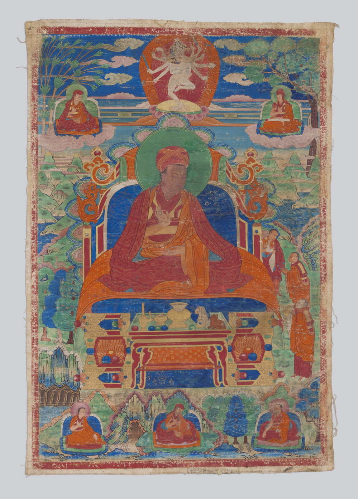 Thangka painting of a red-robed lama seated on an ornate throne, with a small multi-armed dancing deity above in a red aureole, flanked by seated figures on clouds, an altar table below, and additional seated figures at the bottom of the composition.