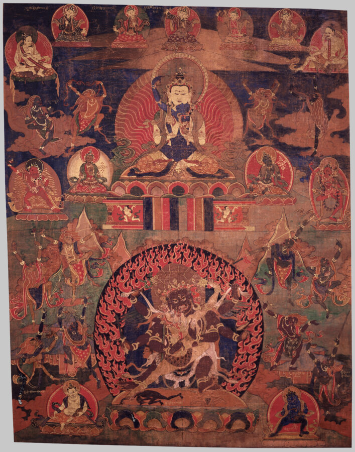 A detailed painting showing multiple deities, with a central seated figure and consort above and a fierce multi-armed deity and consort dancing within a ring of flames below. Other smaller deities in various poses are arranged around the three largest deities.