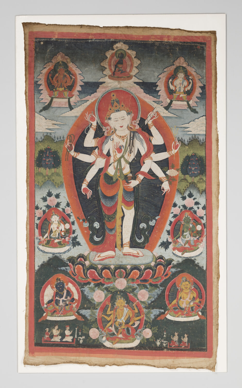 Thangka painting of a standing multi-armed deity with a crown and orange aureole, poised on a lotus base, surrounded by six smaller seated deities in framed roundels and a green landscape background.