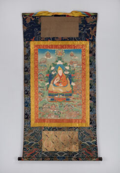 Painting of seated figure with red and saffrom robes surrounded nby smaller figures and buildings mounted on vertical strip of patterned blue fabric.