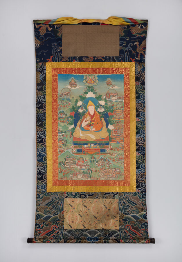 Painting of seated figure with red and saffrom robes surrounded nby smaller figures and buildings mounted on vertical strip of patterned blue fabric.