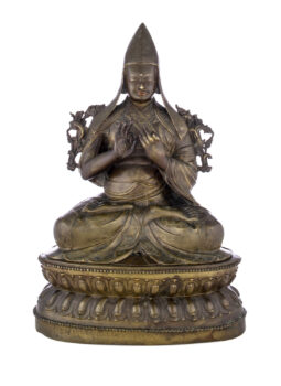 Figure sitting cross legged on a pedestal wearing a pointed hat with long earflaps. Both hands are raised to his chest with index fingers touching thumbs.