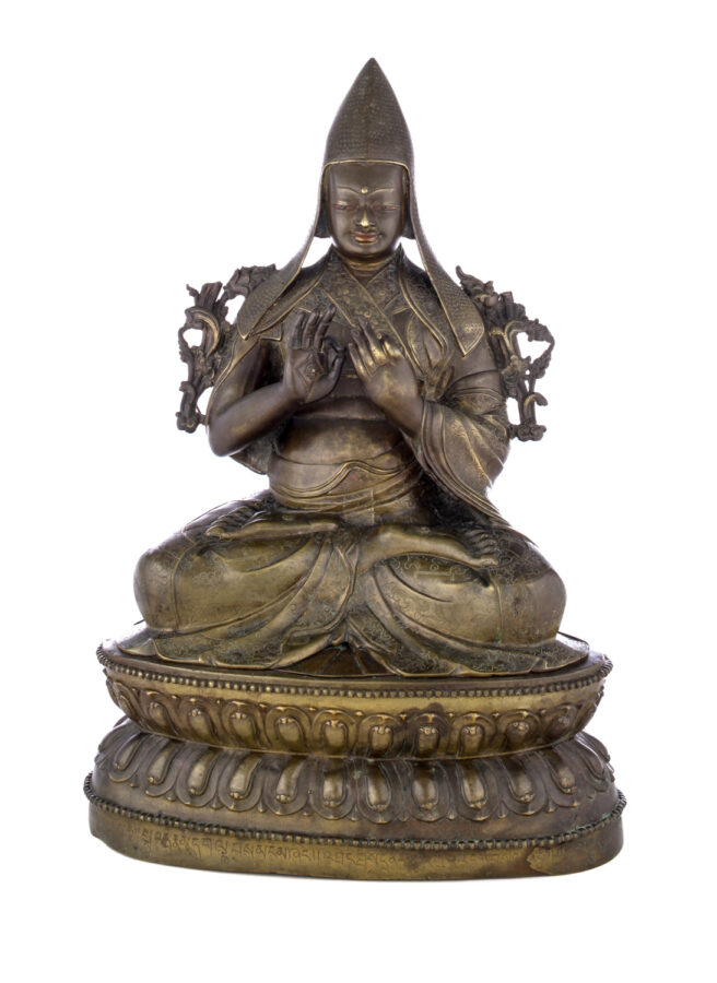 Figure sitting cross legged on a pedestal wearing a pointed hat with long earflaps. Both hands are raised to his chest with index fingers touching thumbs.