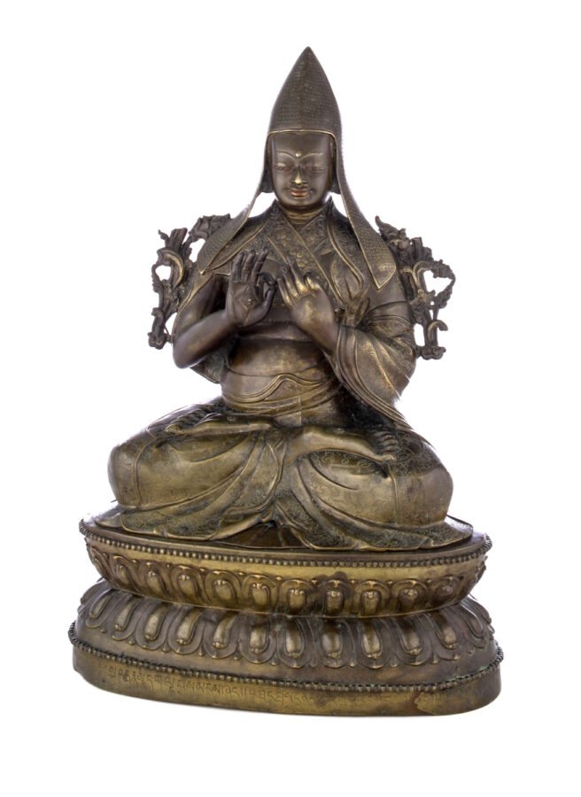 Figure sitting cross legged on a pedestal wearing a pointed hat with long earflaps. Both hands are raised to his chest with index fingers touching thumbs.