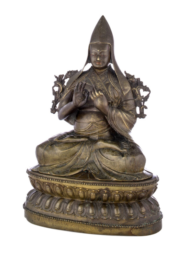 Figure sitting cross legged on a pedestal wearing a pointed hat with long earflaps. Both hands are raised to his chest with index fingers touching thumbs.