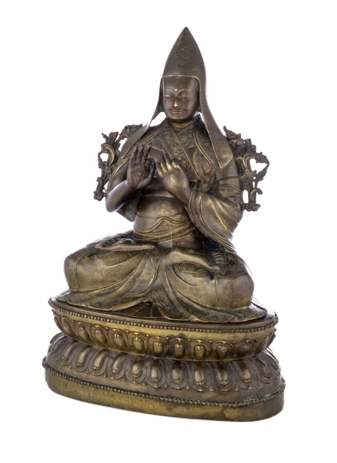 Figure sitting cross legged on a pedestal wearing a pointed hat with long earflaps. Both hands are raised to his chest with index fingers touching thumbs.