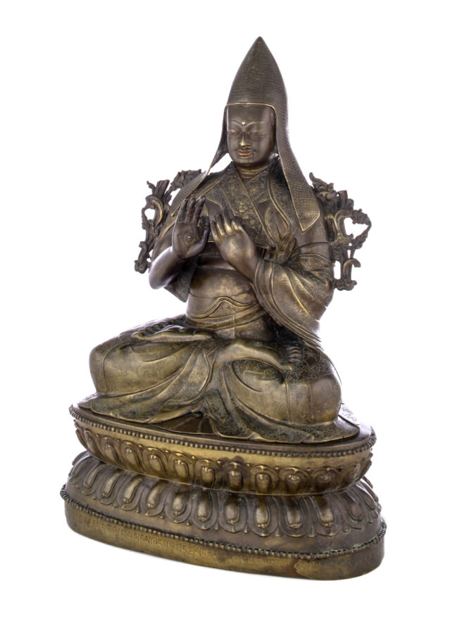 Figure sitting cross legged on a pedestal wearing a pointed hat with long earflaps. Both hands are raised to his chest with index fingers touching thumbs.