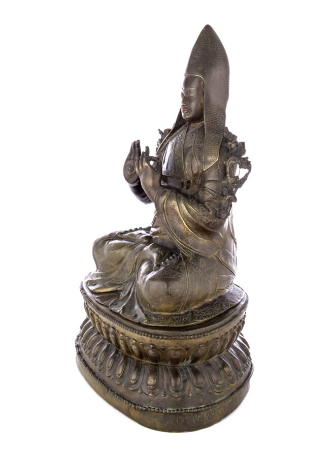 Side view of figure sitting cross legged on a pedestal wearing a pointed hat with long earflaps. Both hands are raised to his chest with index fingers touching thumbs.