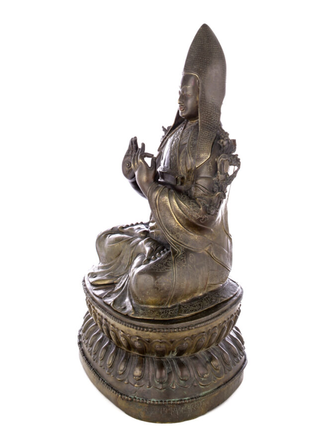 Side view of figure sitting cross legged on a pedestal wearing a pointed hat with long earflaps. Both hands are raised to his chest with index fingers touching thumbs.