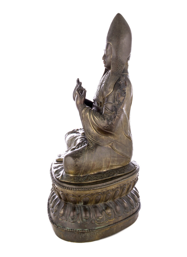 Side view of figure sitting cross legged on a pedestal wearing a pointed hat with long earflaps. Both hands are raised to his chest with index fingers touching thumbs.