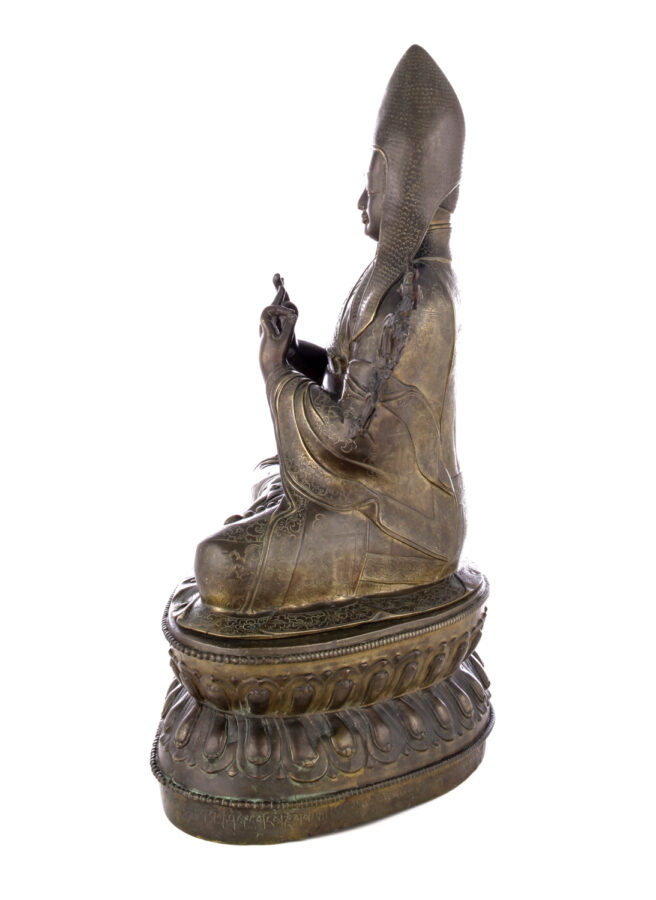 Side view of figure sitting cross legged on a pedestal wearing a pointed hat with long earflaps. Both hands are raised to his chest with index fingers touching thumbs.