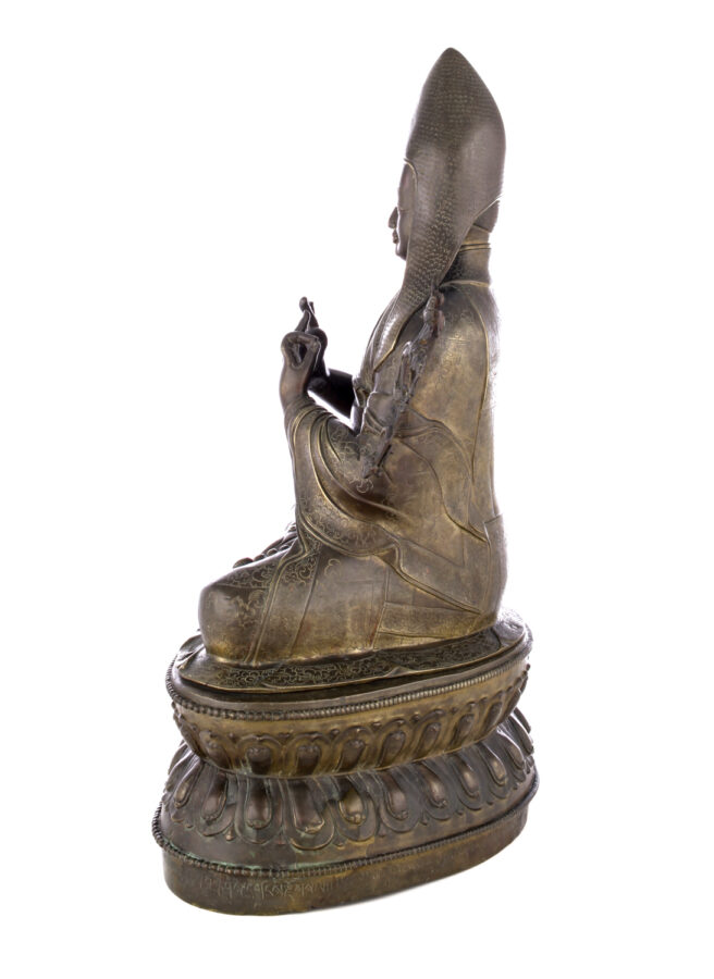 Side view of figure sitting cross legged on a pedestal wearing a pointed hat with long earflaps. Both hands are raised to his chest with index fingers touching thumbs.