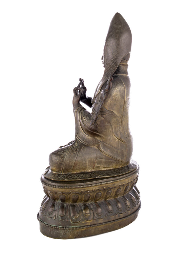 Side view of figure sitting cross legged on a pedestal wearing a pointed hat with long earflaps. Both hands are raised to his chest with index fingers touching thumbs.