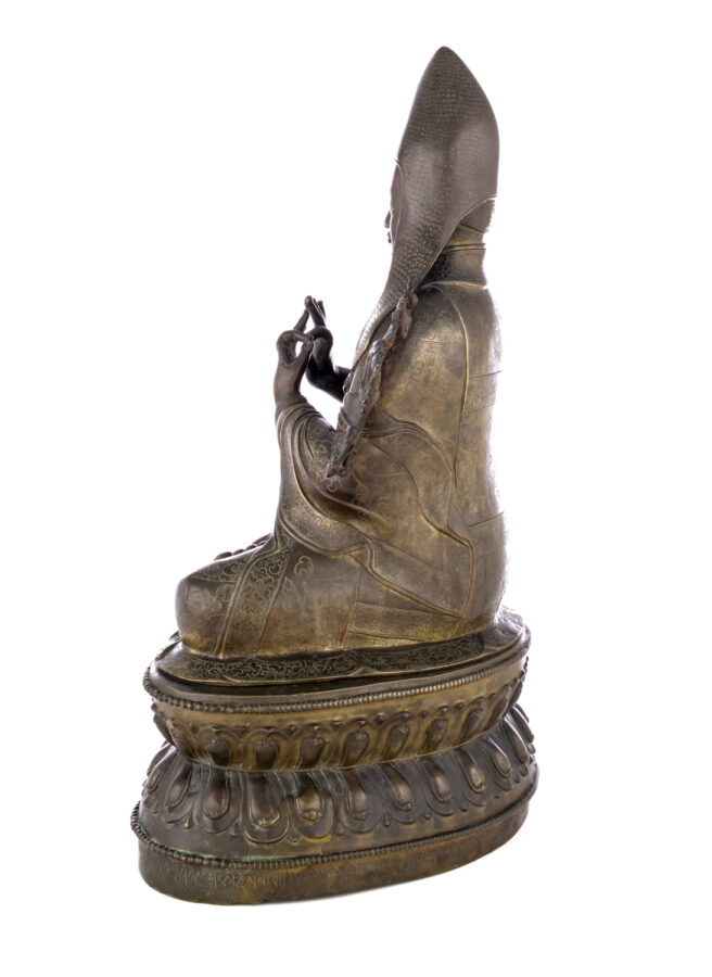 Side view of figure sitting cross legged on a pedestal wearing a pointed hat with long earflaps. Both hands are raised to his chest with index fingers touching thumbs.