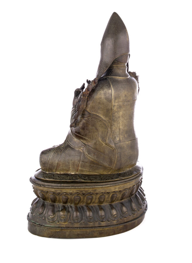 View from behind of figure sitting cross legged on a pedestal wearing a pointed hat with long earflaps.