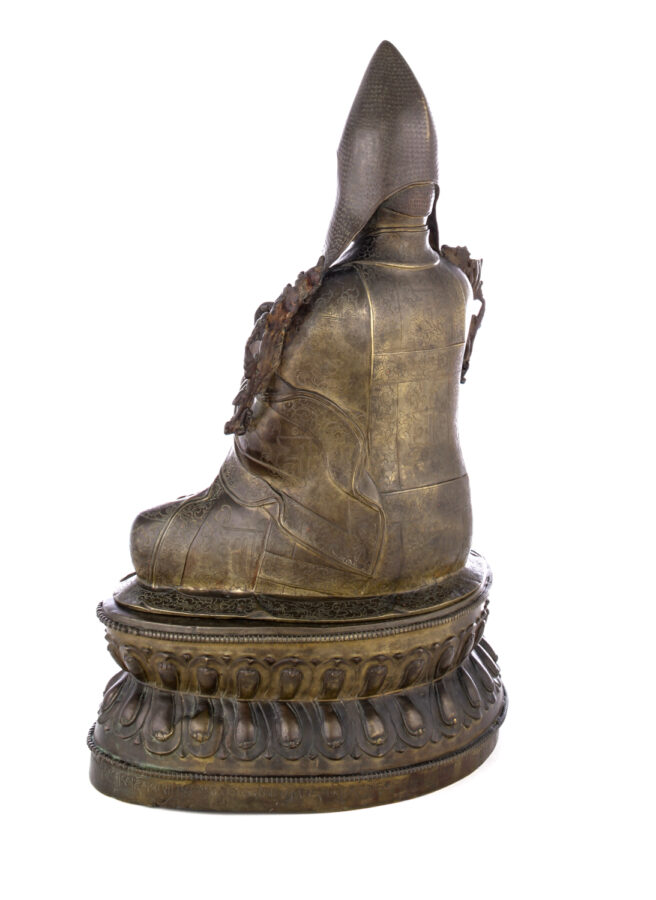 View from behind of figure sitting cross legged on a pedestal wearing a pointed hat with long earflaps.