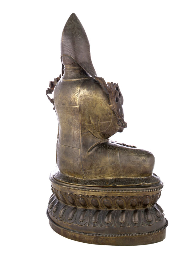 View from behind of figure sitting cross legged on a pedestal wearing a pointed hat with long earflaps.