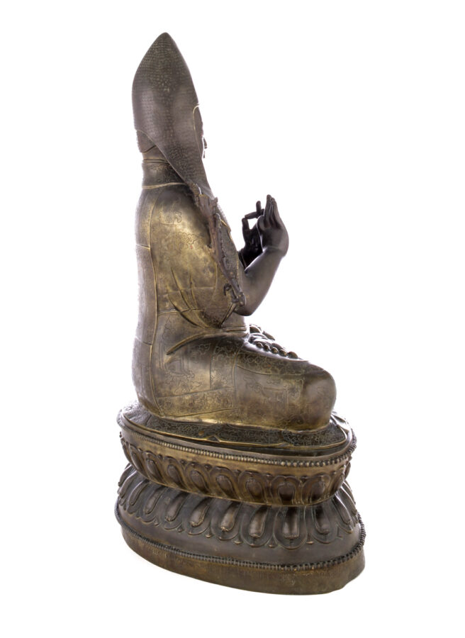 Side view of figure sitting cross legged on a pedestal wearing a pointed hat with long earflaps. Both hands are raised to his chest with index fingers touching thumbs.