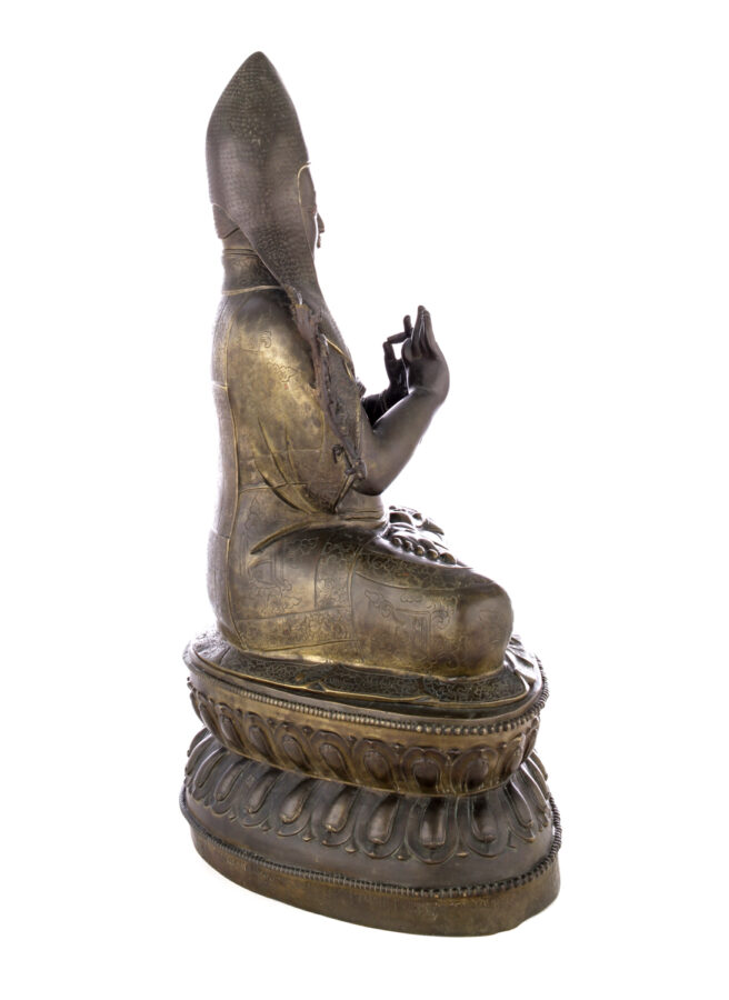 Side view of figure sitting cross legged on a pedestal wearing a pointed hat with long earflaps. Both hands are raised to his chest with index fingers touching thumbs.