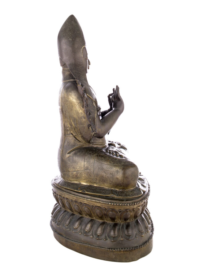 Side view of figure sitting cross legged on a pedestal wearing a pointed hat with long earflaps. Both hands are raised to his chest with index fingers touching thumbs.