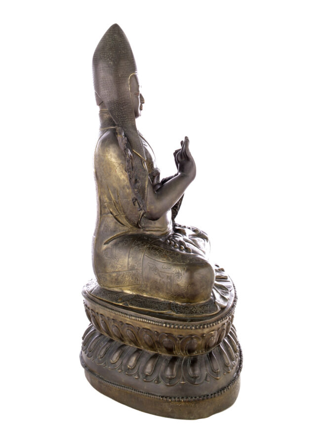 Side view of figure sitting cross legged on a pedestal wearing a pointed hat with long earflaps. Both hands are raised to his chest with index fingers touching thumbs.