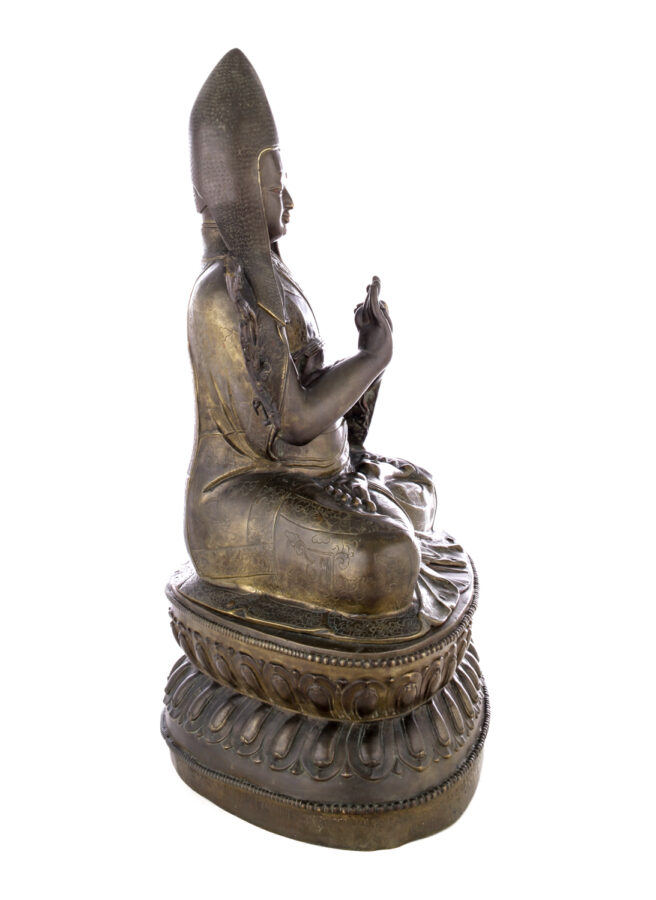 Side view of figure sitting cross legged on a pedestal wearing a pointed hat with long earflaps. Both hands are raised to his chest with index fingers touching thumbs.