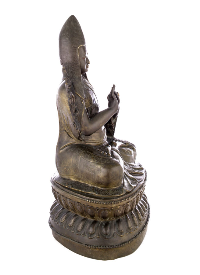 Side view of figure sitting cross legged on a pedestal wearing a pointed hat with long earflaps. Both hands are raised to his chest with index fingers touching thumbs.