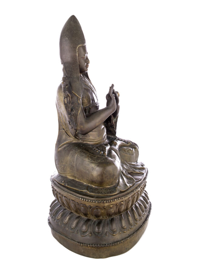 Side view of figure sitting cross legged on a pedestal wearing a pointed hat with long earflaps. Both hands are raised to his chest with index fingers touching thumbs.