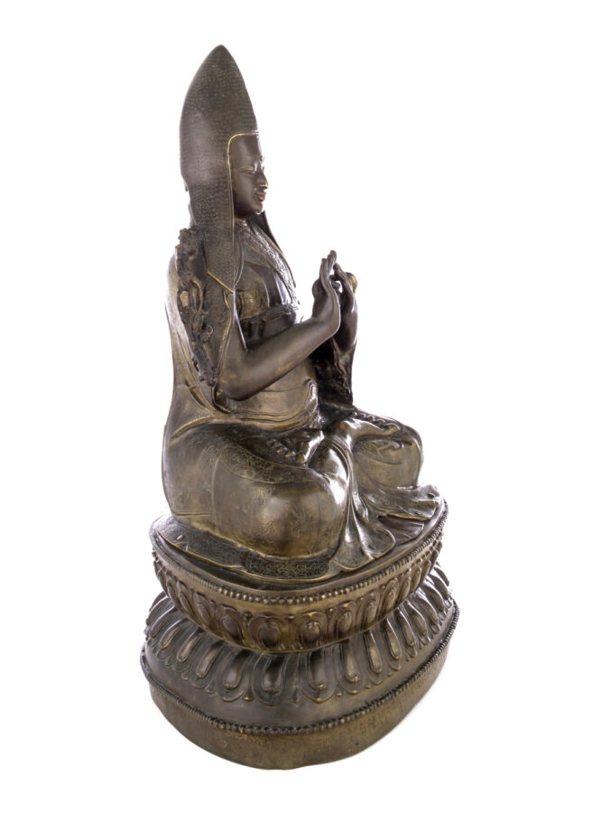 Side view of figure sitting cross legged on a pedestal wearing a pointed hat with long earflaps. Both hands are raised to his chest with index fingers touching thumbs.