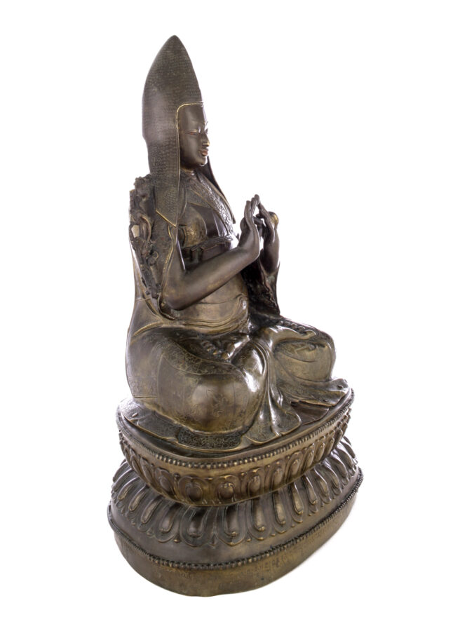 Side view of figure sitting cross legged on a pedestal wearing a pointed hat with long earflaps. Both hands are raised to his chest with index fingers touching thumbs.