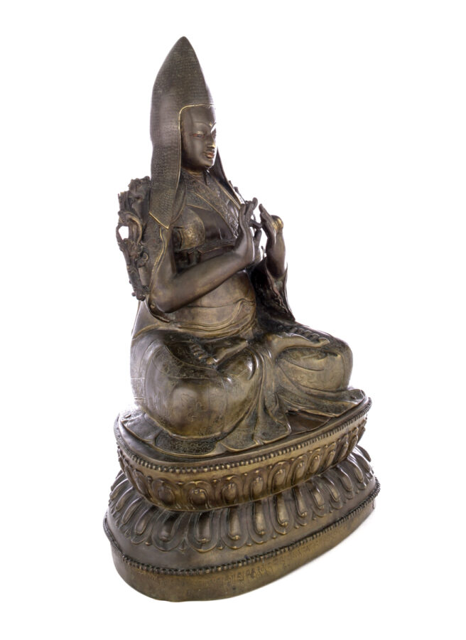 Figure sitting cross legged on a pedestal wearing a pointed hat with long earflaps. Both hands are raised to his chest with index fingers touching thumbs.