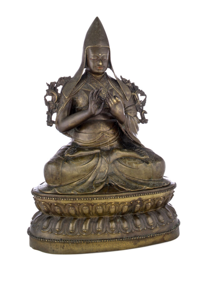 Figure sitting cross legged on a pedestal wearing a pointed hat with long earflaps. Both hands are raised to his chest with index fingers touching thumbs.