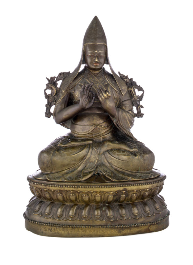 Figure sitting cross legged on a pedestal wearing a pointed hat with long earflaps. Both hands are raised to his chest with index fingers touching thumbs.