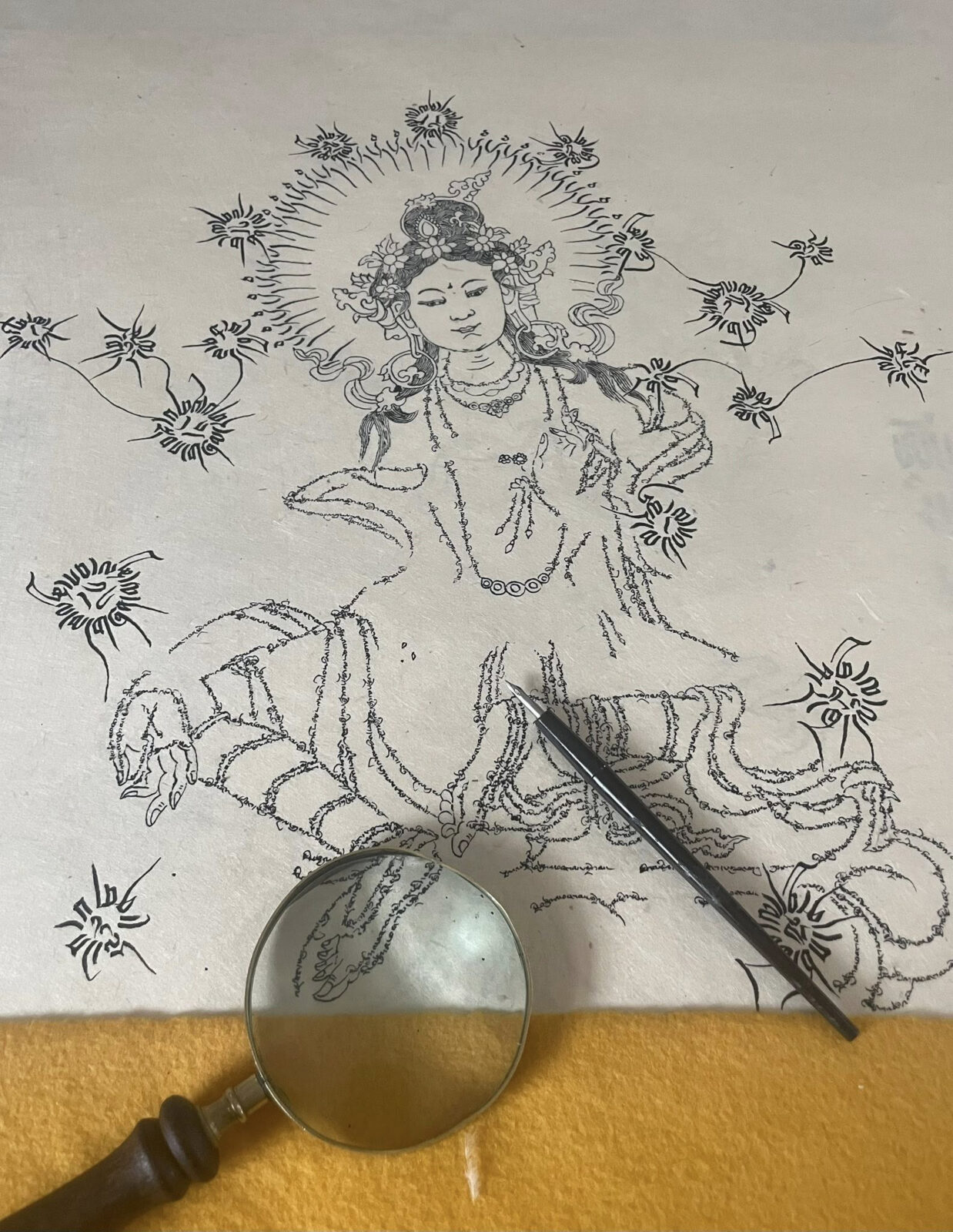 A magnifying glass and calligraphy pen rest on a paper with a drawing of a deity