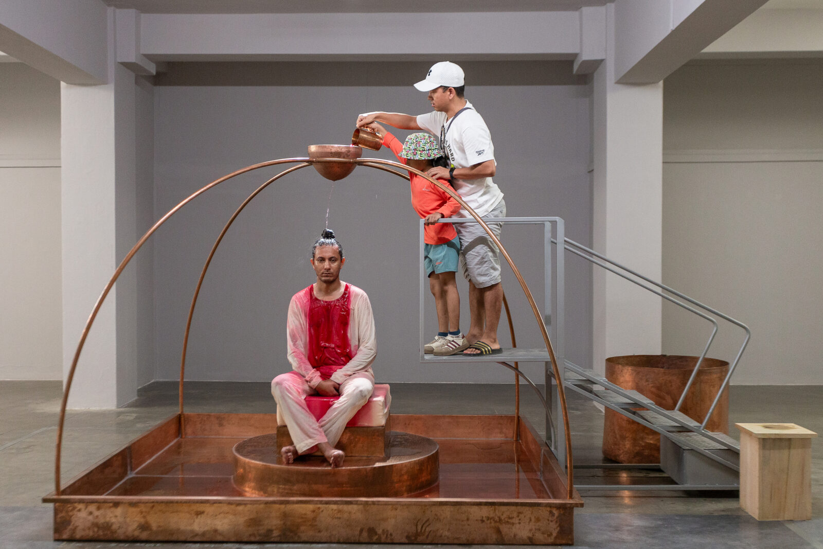 An adult and a child pour liquid into a suspended bronze bowl over a seated man who has red liquid running down his white shirt and pants
