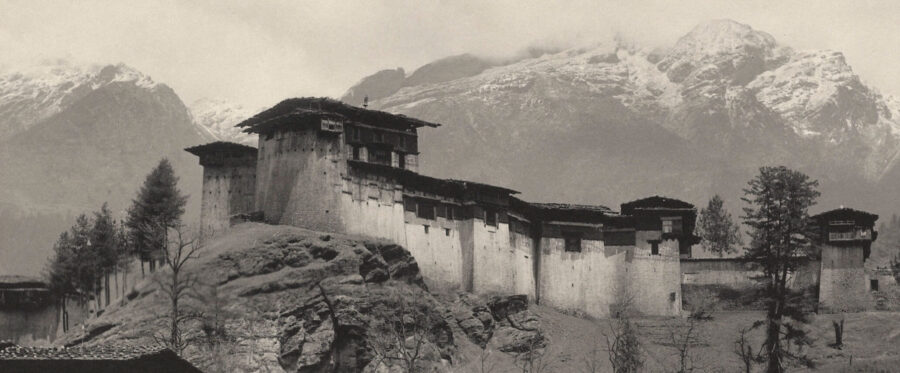 Black-and-white image of long horizontal building on rocky outcropping with mountains in background.