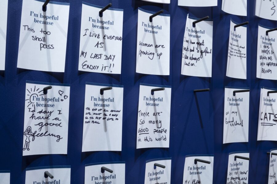 Rows of white cards on pegs printed with "I'm hopeful because" in blue. Answers written in black ink below blue text include "This too shall pass" and "I live everyday like my last day. Enjoy it!!"