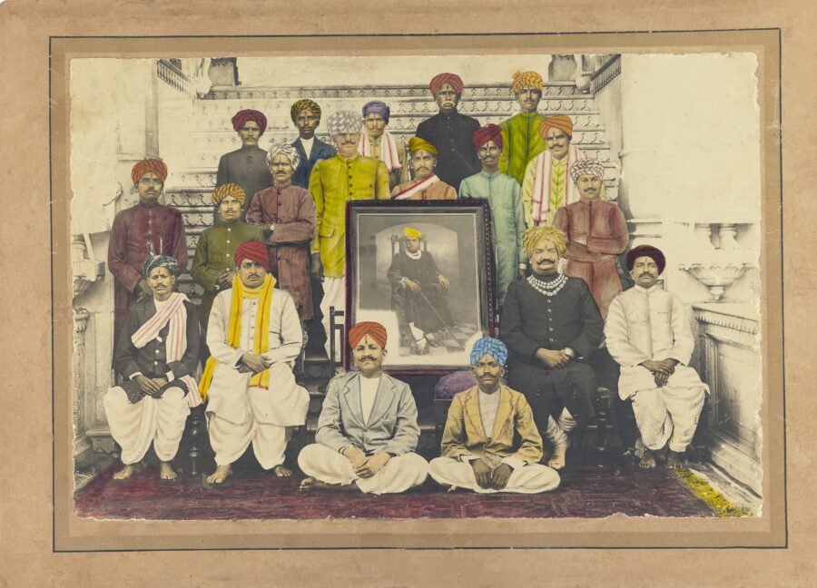 Photograph of men in turbans seating and standing around framed portrait of seated man with yellow turban.