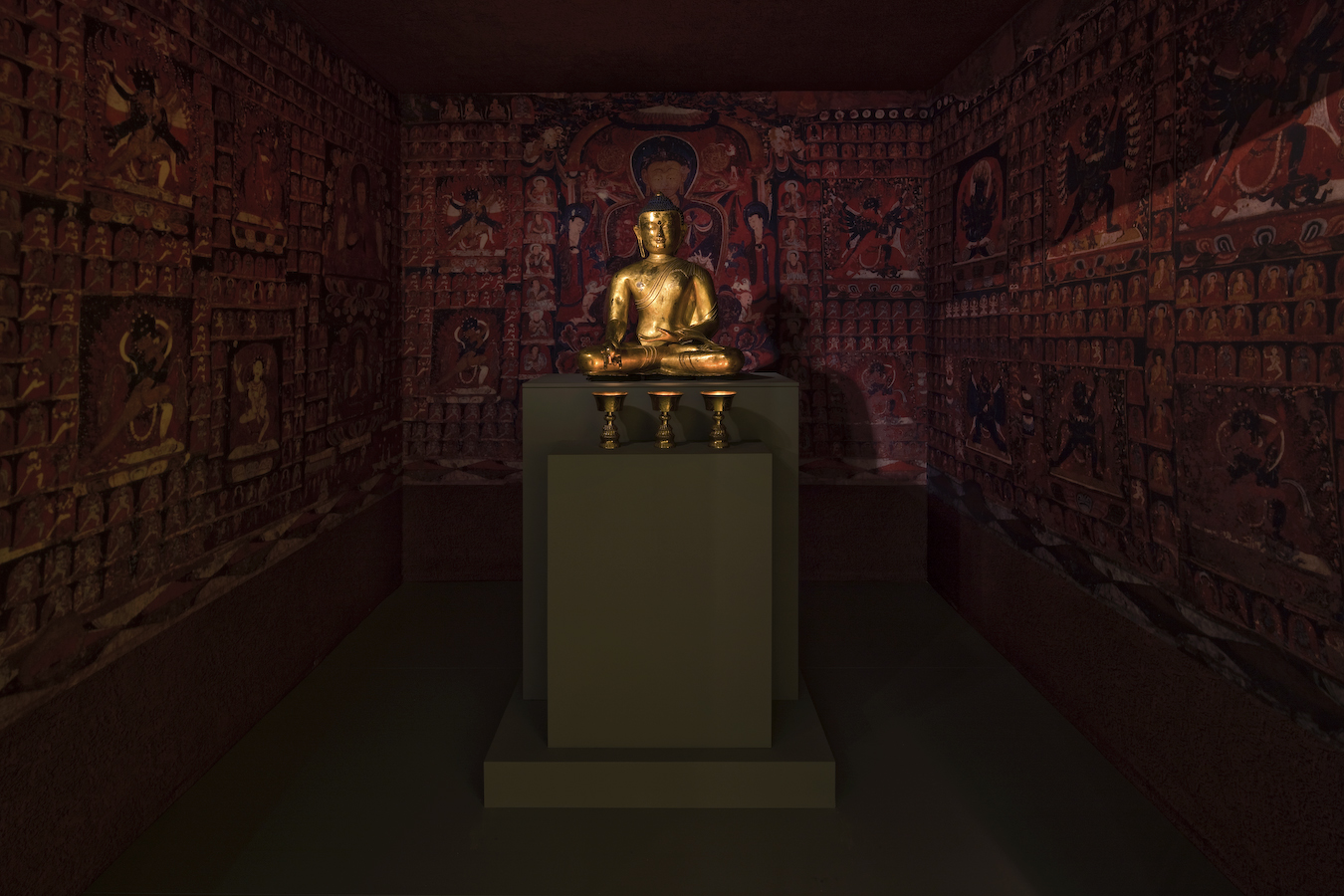 Statue of golden seated figure on pedestal in room surrounded by colorful but paintings of figures and architectural elements.