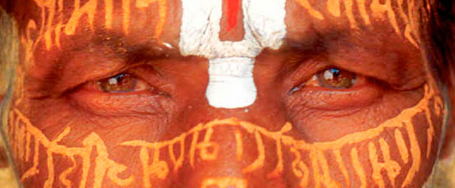 Close-up of human face painted red with white design between eyebrows and yellow designs on nose, cheeks, and forehead.