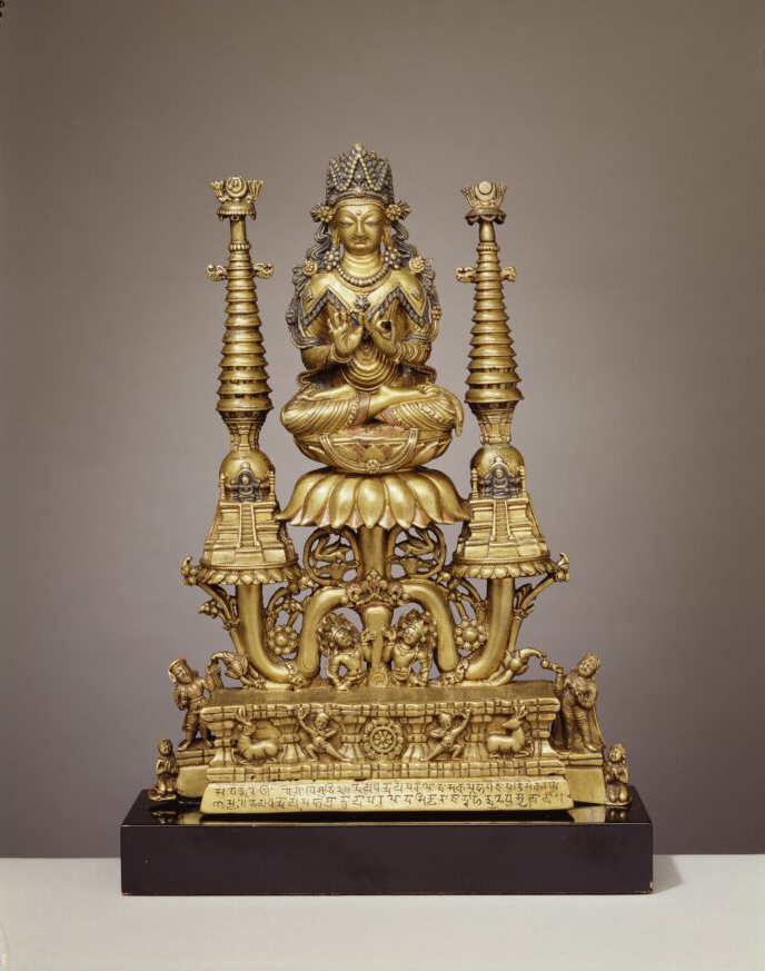 Golden statue of seated figure on lotus blossom flanked by miniature buildings with very tall roofs.