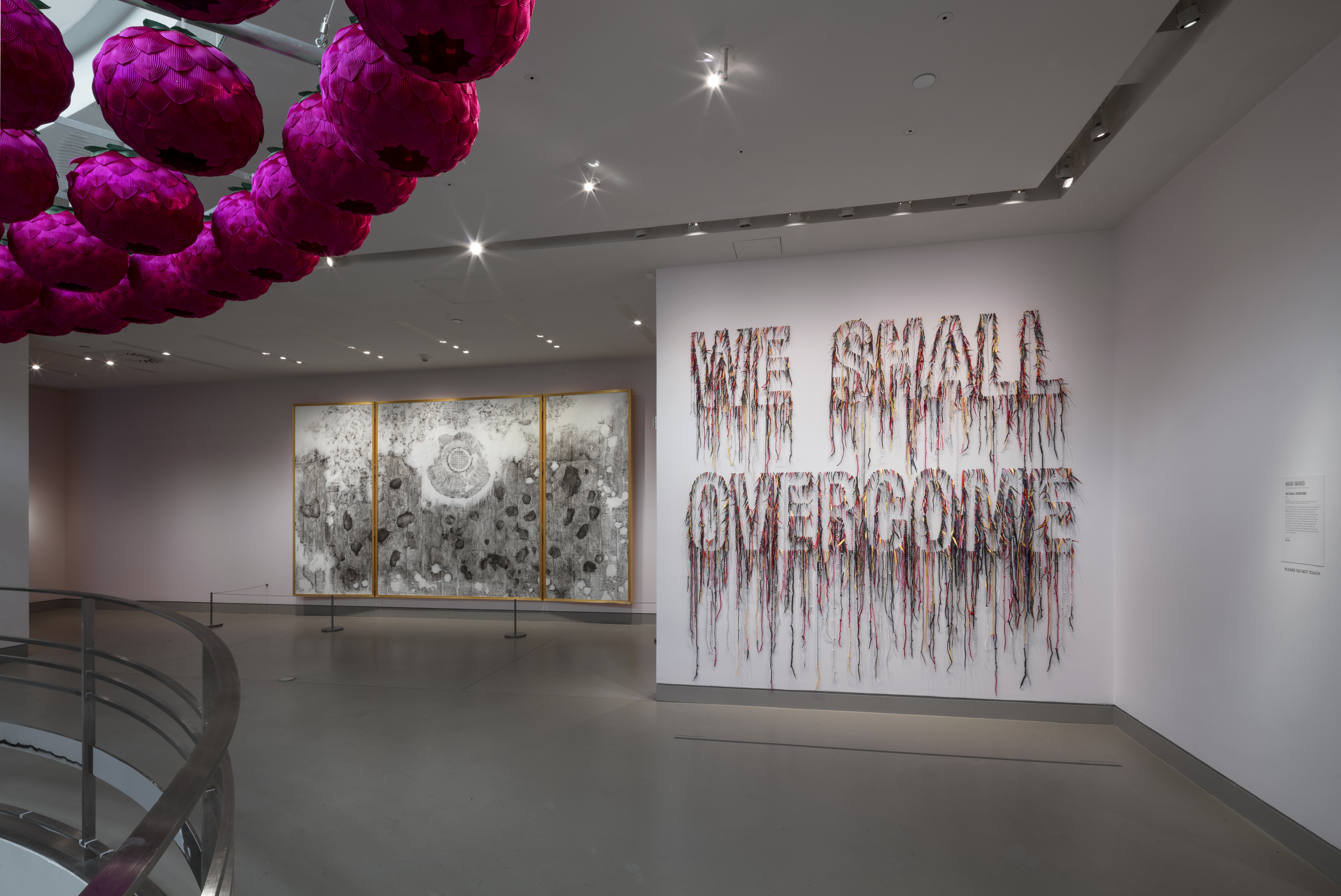 View of exhibition in gallery showing purple sculptural form overhead, large black-and-white framed work on back wall, and multicolored thread spelling "WE SHALL OVERCOME".