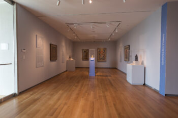 A photograph of an exhibition hall with artworks on the walls and pedestals at the McMullen Museum of Art in Boston