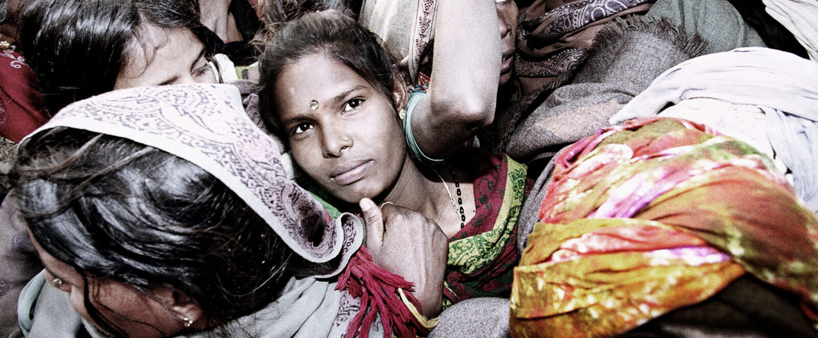 Photograph of woman with jewel above her eyebrows looking up and to the right, surrounded by tightly packed bodies with colorful fabrics.