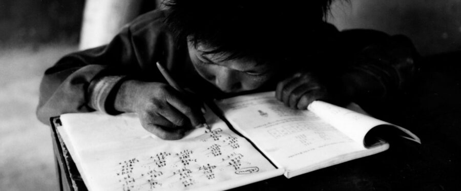 Black-and-white image of child leaning over math book and writing numbers in notebook in ink.