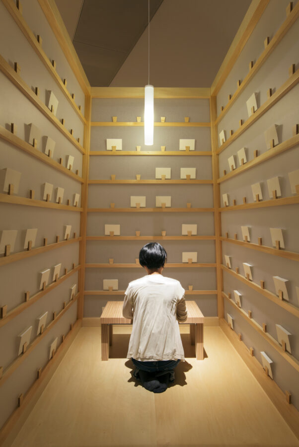 Back of person kneeling at small wooden table, surrounded by three blue walls with rows of wooden rails holding small white boxes.