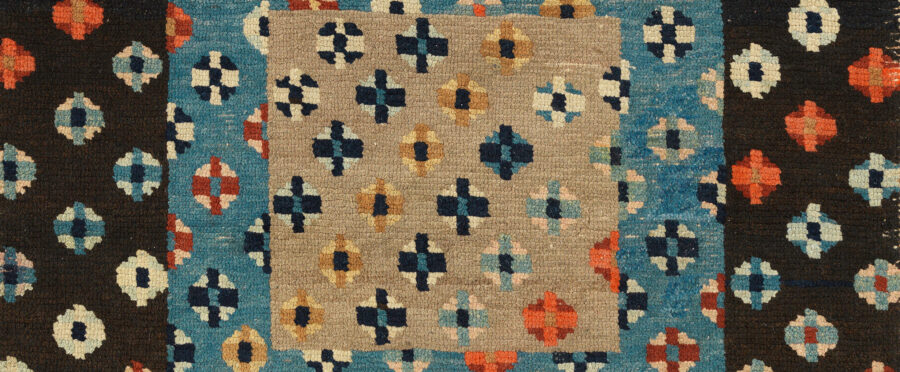 Rug with geometric diamond shapes of dark blue, light blue, cream, red, and orange on larger squares of cream and blue.