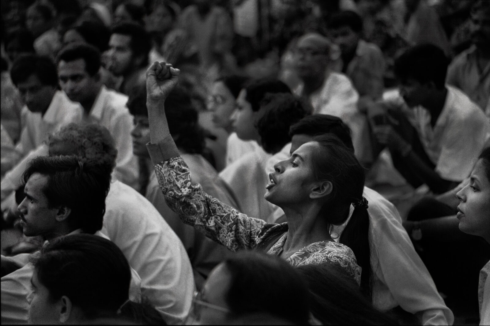 Black-and-white image of woman with raised fist seated among many people.