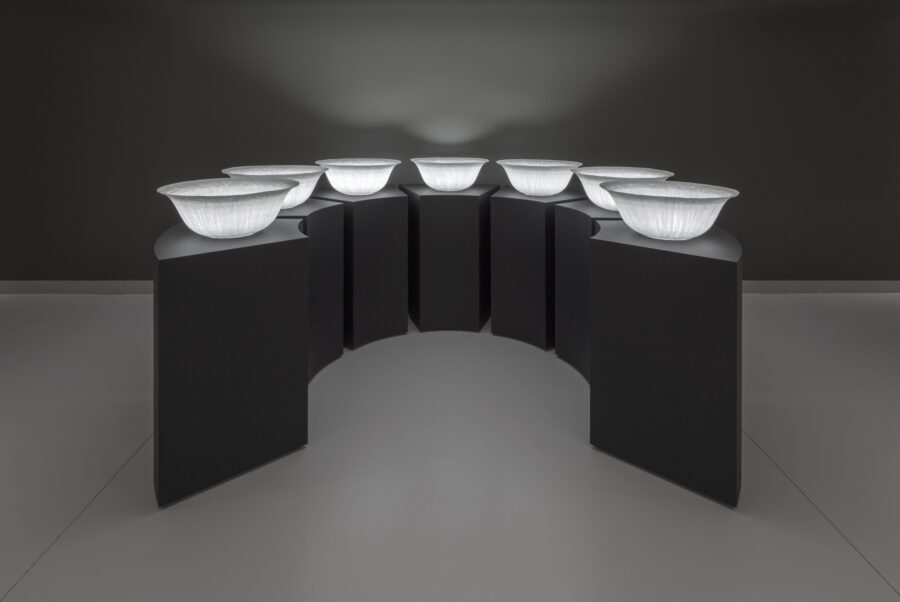 Seven glowing white bowls, each on its own black pedestal, in darkened gallery.